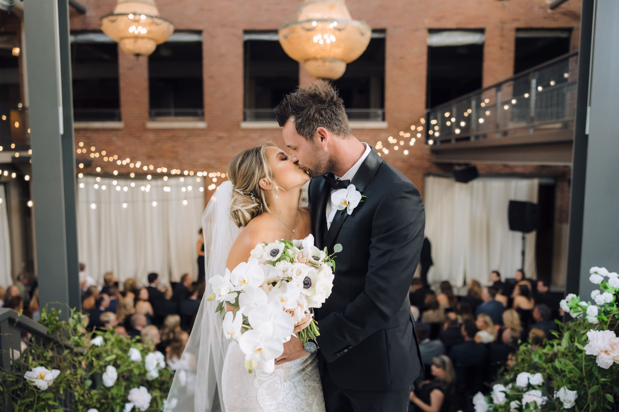 Bride and groom sharing first kiss during Chicago wedding ceremony with white floral arrangements and string lights designed by Erin McDonald