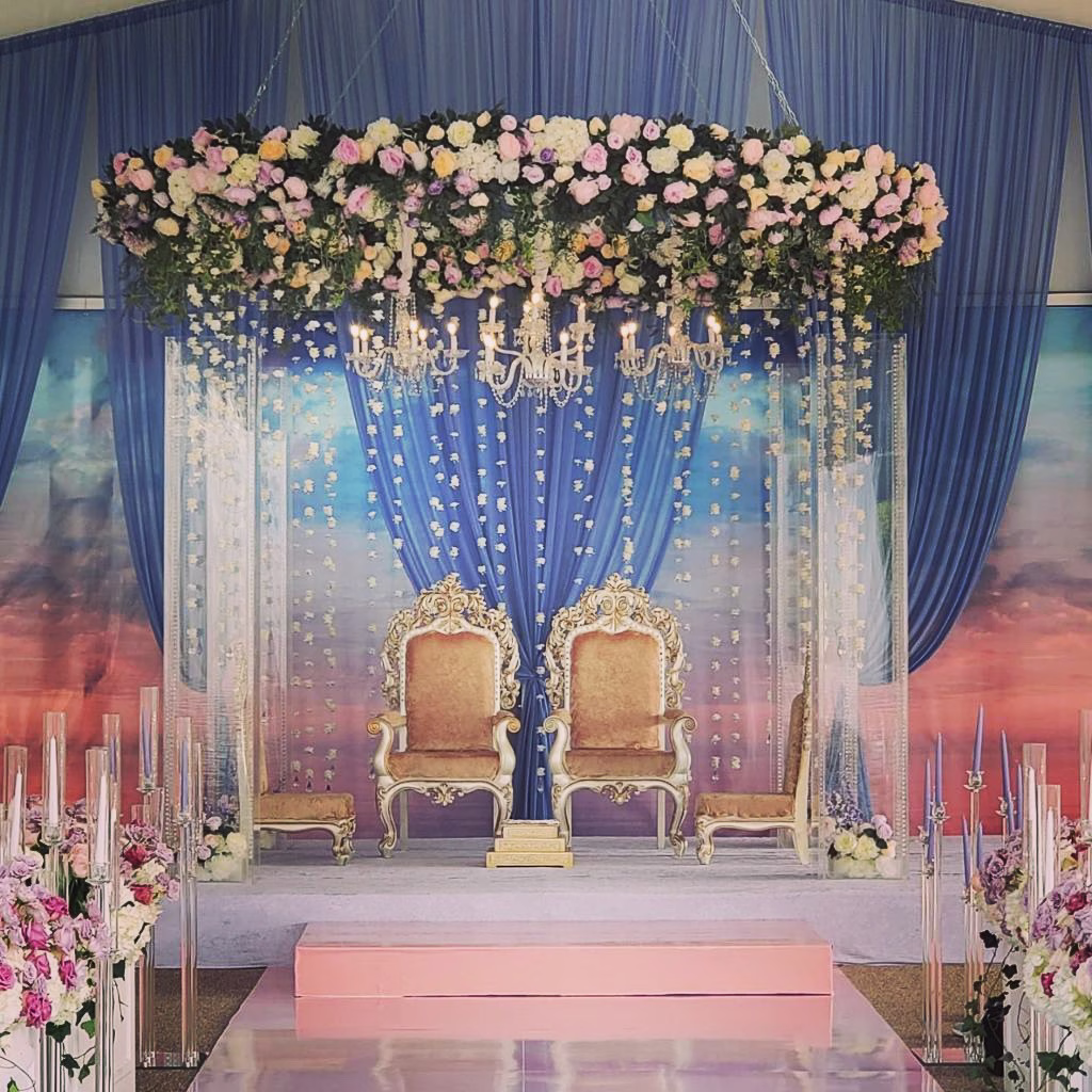 Elegant South Asian wedding ceremony setup with ornate golden chairs, crystal chandelier, cascading floral ceiling installation and blue draping designed by Erin McDonald