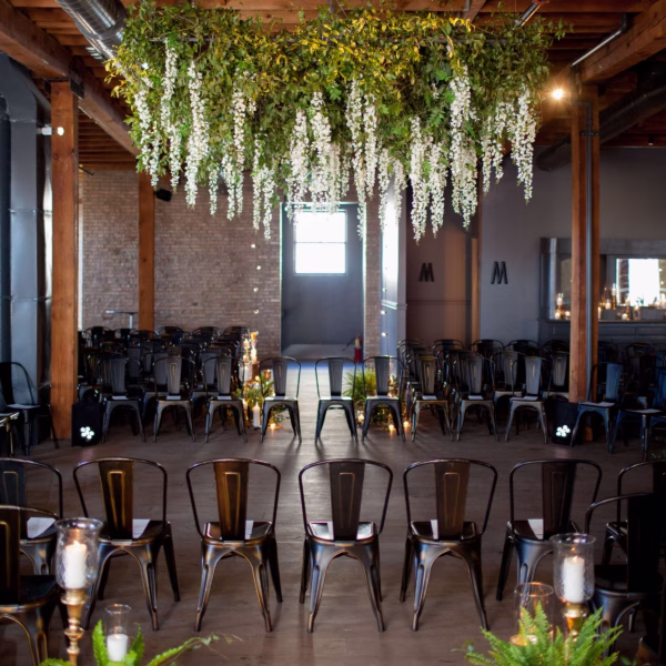 Industrial wedding ceremony space with dramatic hanging white wisteria floral installation suspended from exposed wooden beams, featuring black metal chairs and candlelit aisle designed by Chicago wedding designer Erin McDonald