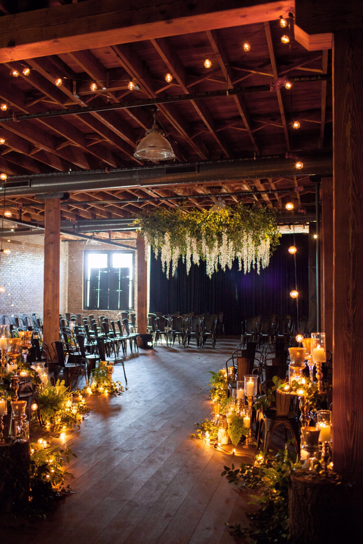 Industrial wedding ceremony space with exposed wooden beams, string lights, hanging floral installation, and candlelit aisle designed by Chicago and Rochester NY wedding designer Erin McDonald